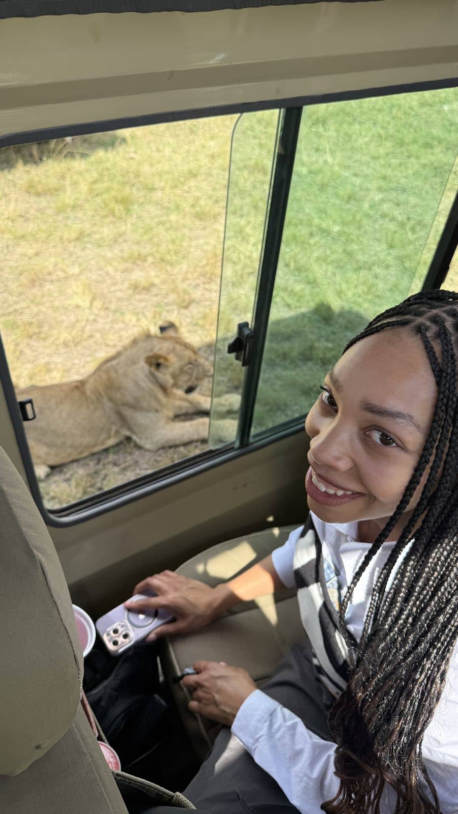 A traveller smiling in the safari vehicle as a lioness rests just outside the window