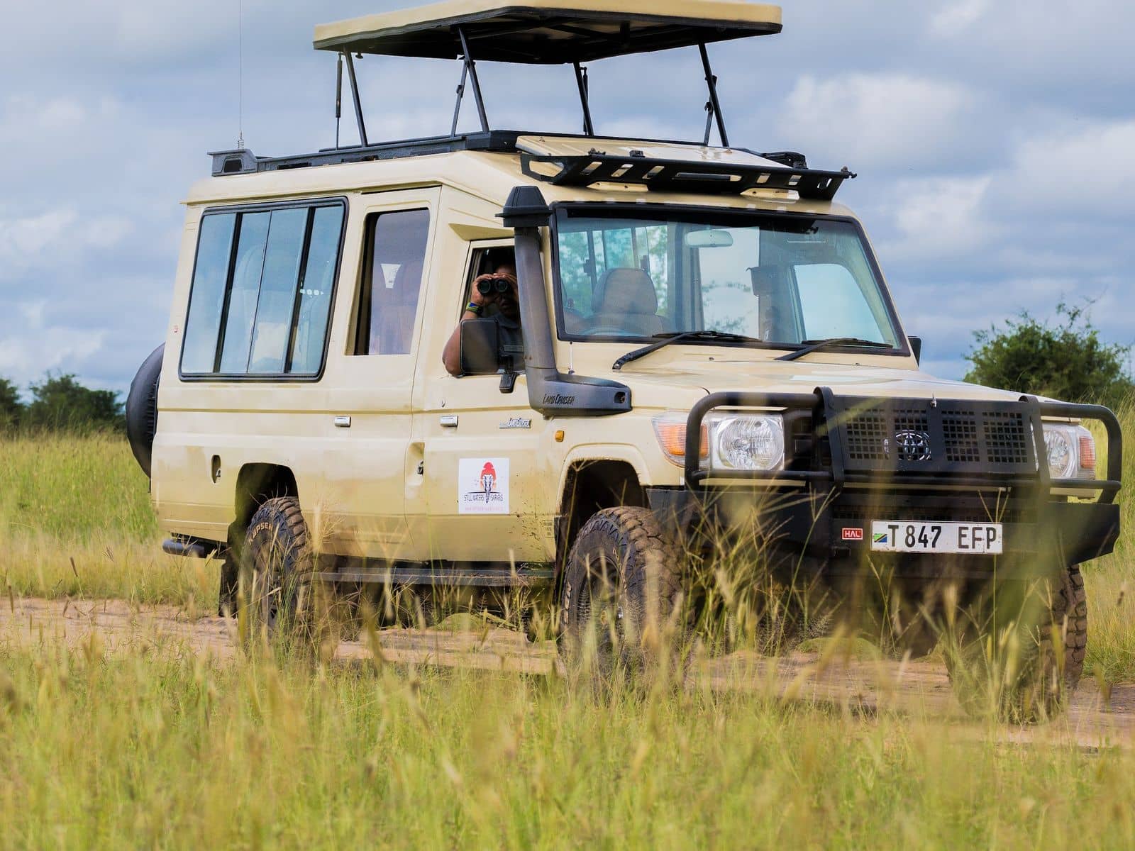 A Still Waters Safaris Land Cruiser with its roof raised on a Serengeti track, the guide scanning the plains through binoculars